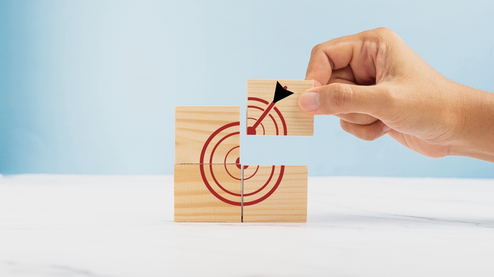 A hand placing a small wooden block with an arrow onto a stack of wooden blocks forming a target symbol, against a light blue background.