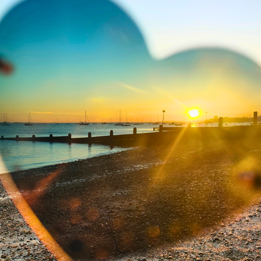 A serene sunset view at Southend-on-Sea beach, featuring wooden groynes stretching into calm waters. The sky transitions from turquoise to golden yellow, with silhouettes of sailboats anchored along the horizon. Sun rays create lens flares across the pebbly beach, while the low tide reveals textured shoreline. The composition captures the peaceful evening atmosphere of the Essex coast.