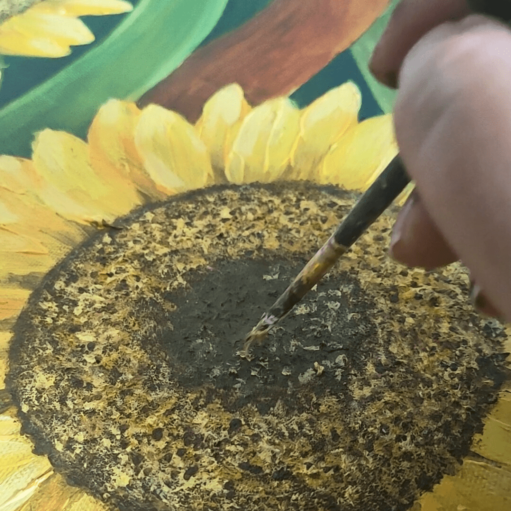 Extreme close-up of a sunflower center in an acrylic painting, showing intricate seed pattern details with rich texture and subtle color variations. A fine brush adds final touches to the complex spiral pattern against bright yellow petals.