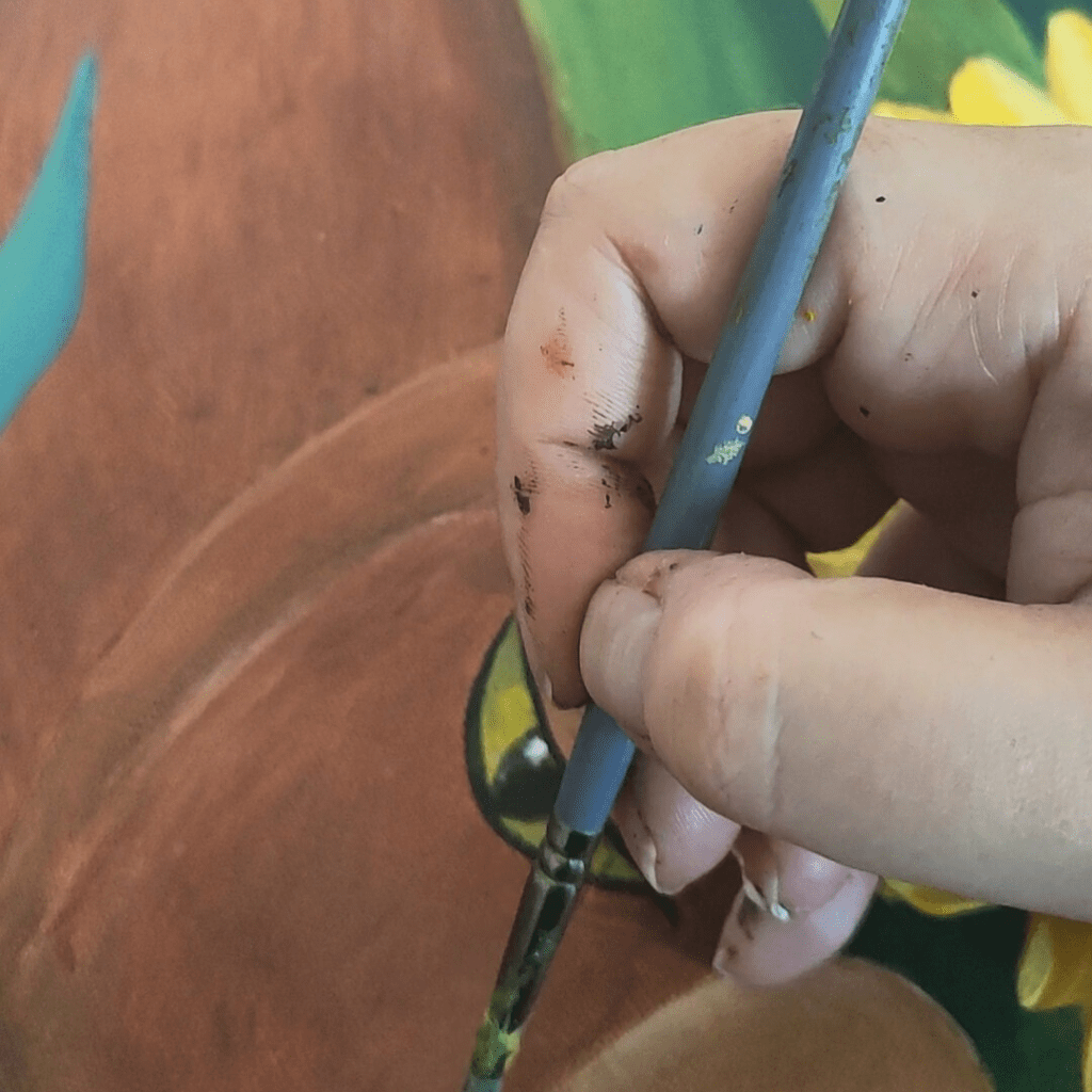 Close-up of an artist's paint-stained fingers holding a brush against a painting background showing terracotta browns and sunny yellows with green foliage.
