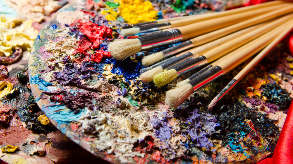 A close-up macro photograph of well-used paint brushes resting on an artist's palette. The palette shows rich textures of dried and fresh acrylic paint in various colors including vibrant red, royal blue, purple, yellow, and turquoise. The brushes are arranged diagonally across the image, with their natural bristles showing signs of use. The wooden handles create a warm contrast against the colorful paint textures.