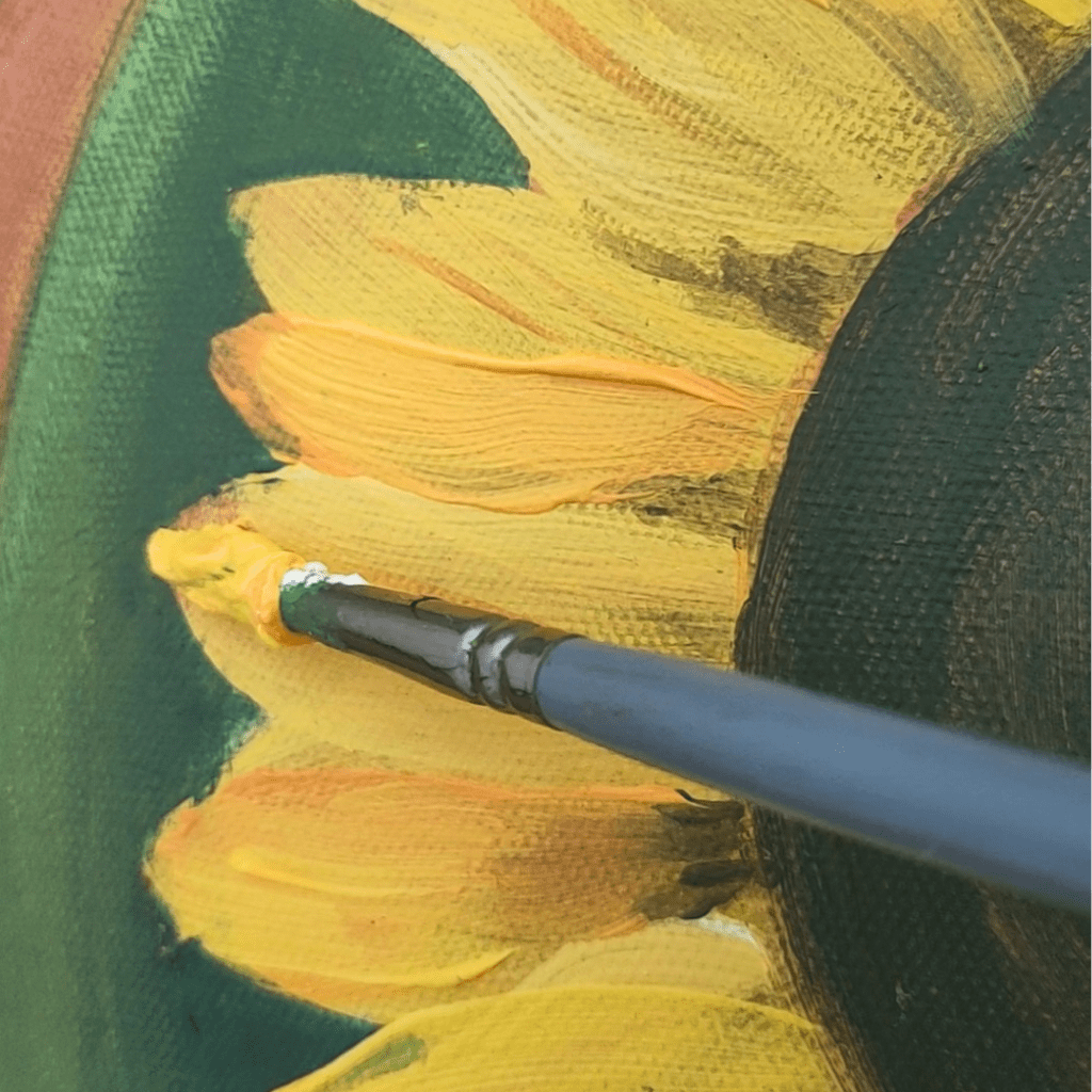 Close-up of an acrylic painting in progress, showing a brush adding detailed strokes to bright yellow sunflower petals against a deep green background. The brush strokes show clear texture and directional movement.