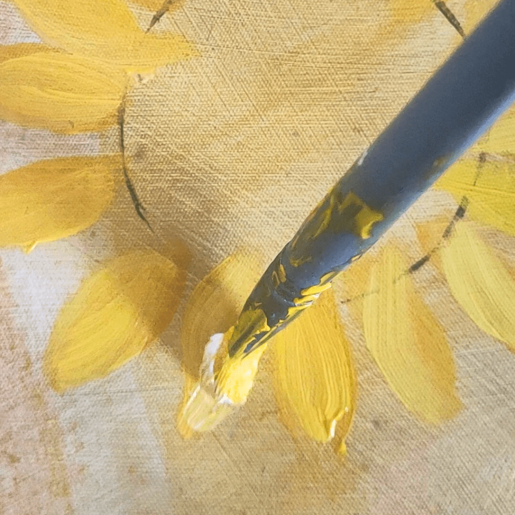Close-up macro photograph of an acrylic painting in progress, showing a paintbrush adding yellow petals to a sunflower against raw canvas texture. The brush has traces of yellow paint, demonstrating the active painting process.