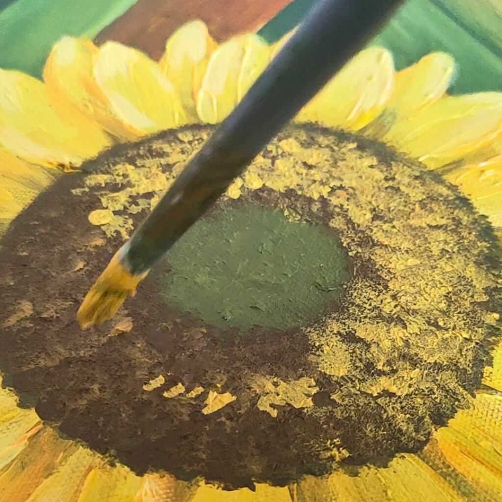 Extreme close-up of an acrylic painting in progress, showing a brush adding textured details to a sunflower's dark brown center, surrounded by bright yellow petals. The painting captures the intricate spiral pattern characteristic of sunflower seeds.
