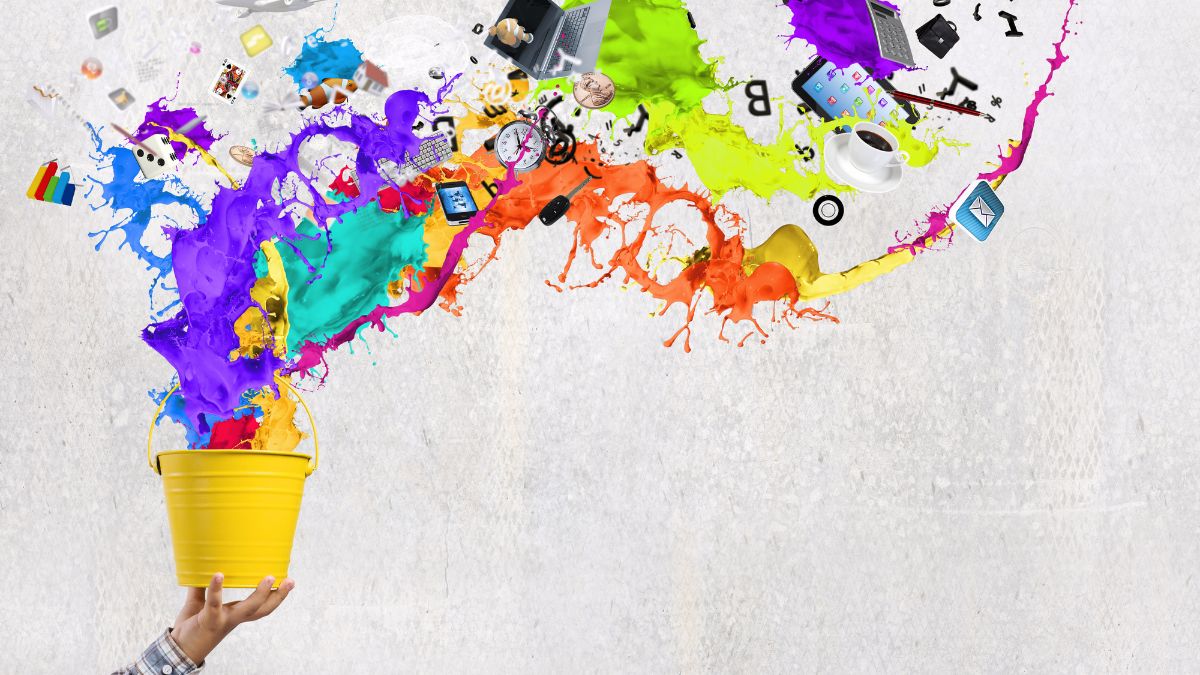A hand holding a yellow bucket with colourful paint splashes bursting out across a light grey background, mixed with small floating icons like a laptop, coffee cup, and digital symbols.