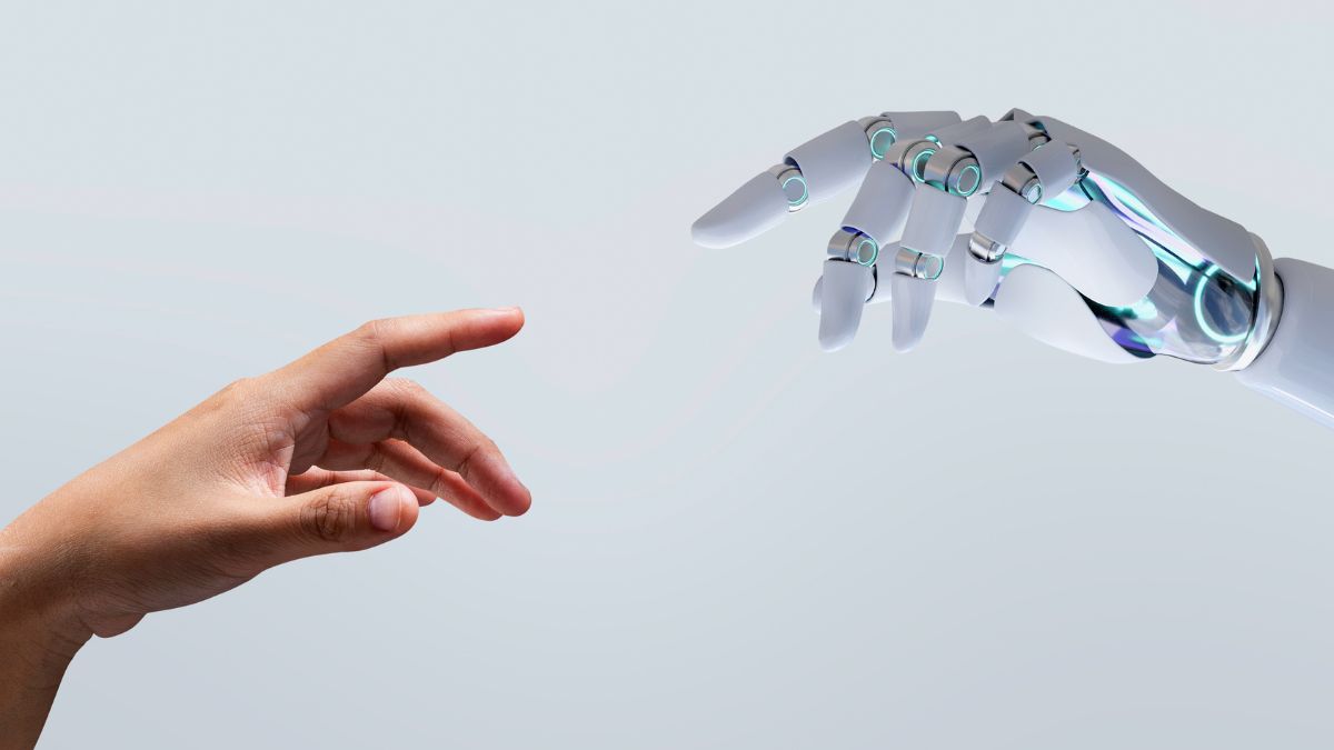 Close-up image of a human hand reaching toward a robotic hand against a light grey background, with both index fingers extended as if about to touch.