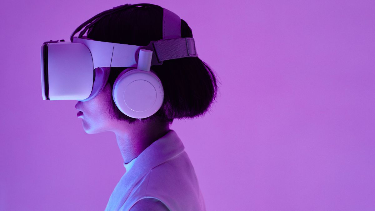 Side profile of a person wearing a VR headset and large headphones against a purple background, lit with pink and violet tones.