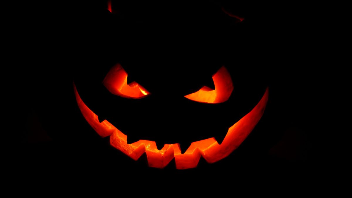 Close-up of a glowing jack-o’-lantern face in darkness, with bright orange light shining through carved eyes and a jagged smiling mouth.